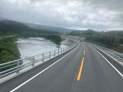 The image depicts a curved asphalt road with yellow and white lane markings, bordered by a metal guardrail. The road is elevated over a body of water, possibly a river, with lush green trees lining the banks. In the background, a mountainous landscape with sparse vegetation and a cloudy sky is visible. There is a single vehicle, a dark SUV, on the road ahead. The overall scene suggests a natural, scenic environment, likely in a rural or mountainous region. The location is identified as Gluggvasshaug, Norway.