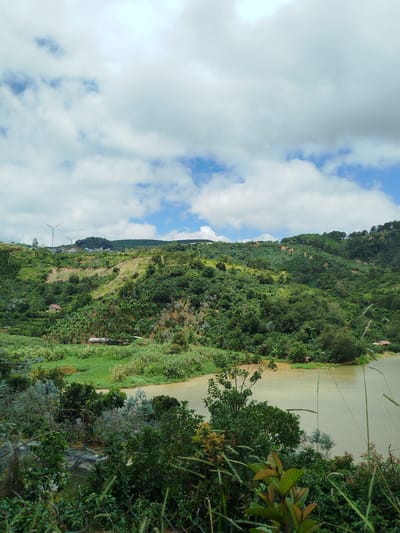 This is an exterior, eye-level, wide shot of a lush green hillside overlooking a calm body of water, likely a lake or river, in Xuân Sơn, Vietnam. The sky is partly cloudy with bright white clouds scattered across a pale blue expanse. The hillside is densely covered in a variety of vegetation, including trees, bushes, and what appear to be agricultural terraces with crops. In the mid-ground, there are a few small structures, including a house with a reddish-brown roof and some farm buildings. On the crest of the hill, two white wind turbines are visible, suggesting a renewable energy installation. The water in the foreground has a murky, greenish-brown hue and reflects the cloudy sky. The overall scene depicts a rural landscape with elements of both nature and human activity.