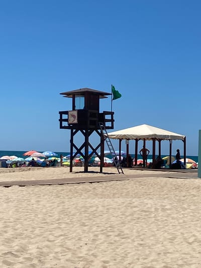 The image depicts a crowded beach scene on a bright, sunny day in Conil de la Frontera, Spain. In the foreground, a wide expanse of light brown sand is visible, with subtle ripples indicating the gentle movement of wind or water. A wooden walkway extends across the sand, leading towards the midground.
Dominating the midground is a dark brown wooden lifeguard tower, with a small enclosed cabin at the top. A ladder is attached to its side, leading up to the cabin. A green flag is flying from a pole on top of the tower. To the right of the lifeguard tower, a large, open-sided, white-covered structure with multiple posts provides shade. Several people are seen in silhouette under this structure, some appearing to be seated or standing.
Further in the background, a vibrant array of colorful beach umbrellas are spread across the sandy area, with many beachgoers seated or lounging beneath them. The sea is visible in the distance, with a clear blue sky above. The overall impression is one of a busy, recreational beach environment on a summer day.