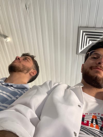 The image displays a low-angle perspective of two adult males indoors, under a white paneled ceiling. The male on the left, visible from the chest up, has a beard and short dark hair, wearing a light blue and white striped polo shirt. He is looking upwards and to his left. The male on the right, also visible from the chest up, has a beard and wears gold-rimmed glasses. He is wearing a white long-sleeved jacket with snap buttons over a white t-shirt with a colorful graphic print. He is generally facing forward. The ceiling features vertical white panels, a circular white light fixture on the upper left, and a square grey ventilation grill on the upper right. A slight indentation is present on one ceiling panel in the upper left. The scene is located in قرية الضبية, Syria.