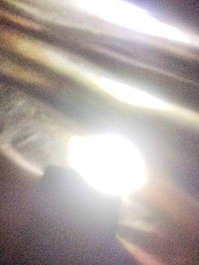 The image presents an abstract composition of light and shadow. Bright, elongated streaks of light dominate the frame, contrasting with darker, blurred areas. A central, intensely bright region appears almost circular, radiating light outwards. The overall effect is ethereal and unclear, with no easily identifiable objects or forms discernible. The texture appears grainy and somewhat obscured.