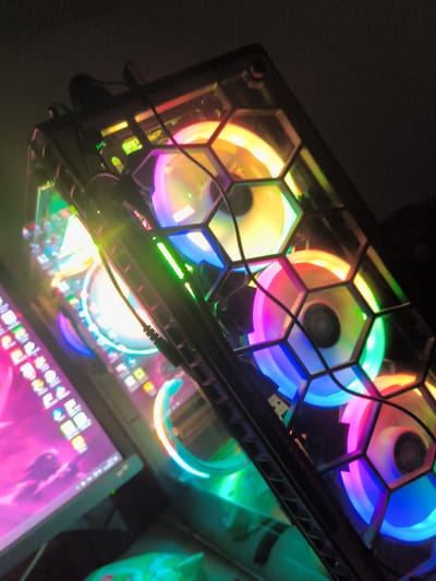 A dimly lit, close-up photograph shows the side of a computer tower with a honeycomb-patterned grill, behind which multi-colored LED cooling fans are visible. To the left of the tower is a computer monitor displaying an interface with icons and a wallpaper that appears to be primarily pink. Some cables and other items, out of focus, are visible in the foreground.
There is no indication that the image was taken in the city of Havixbeck, Germany.