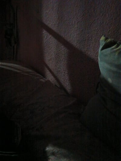 The image depicts an interior scene, likely a bedroom, with a strong emphasis on shadows and texture. A bed or sofa occupies the majority of the foreground, covered in dark fabric that appears to be wrinkled or folded. A diagonal shadow from a window or an object casts across the textured wall in the background, which has a rough, speckled appearance. To the right of the frame, a portion of a green pillow or cushion is visible, its fabric bunched up. The lighting suggests it is either early morning or late evening, with limited ambient light. The overall atmosphere is dim and somewhat stark. There is no indication of the city of Juma, Uzbekistan within the image itself, nor any people or specific actions taking place.