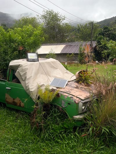 A weathered green pickup truck, covered with a light-colored tarp, sits abandoned amidst overgrown vegetation in Ban Ang Ka Noi, Thailand. A large LED floodlight and a smaller solar panel rest on the tarp-covered hood. The truck's paint is peeling, revealing rust underneath, and the surrounding area is dense with tall grasses and various plants. In the background, a structure with a corrugated metal roof is visible, partially obscured by trees and foliage. Rolling hills, shrouded in mist, form the distant landscape under a cloudy sky. The scene conveys a sense of neglect and nature reclaiming an industrial object.