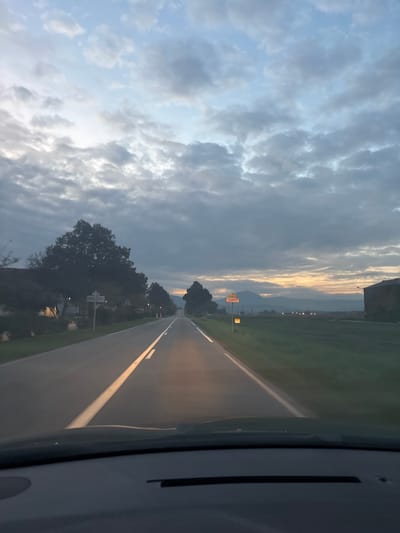 The image depicts a straight two-lane paved road, likely a main route, at twilight (sunrise or sunset) in Ebersheim, France. The viewpoint is from inside a vehicle, looking forward through the windshield, with the vehicle's dashboard visible at the bottom of the frame. The road is marked with double solid white lines in the center and white lines on the shoulders, illuminated by the vehicle's headlights.
On the left side of the road, a line of deciduous trees and several residential or village structures are visible, along with a white, multi-armed directional signpost. On the right, a grassy field borders the road, extending towards a large, dark, rectangular building in the mid-distance. A prominent red and yellow road sign, identifying the location as "Ebersheim," stands on the right shoulder, with a smaller yellow sign positioned below it. In the far distance, a chain of mountains outlines the horizon, where a band of warm orange and yellow light suggests the sun's position. The sky is largely overcast with multiple layers of grey and white clouds, transitioning to lighter blue tones at the top. No people or other vehicles are distinctly visible.