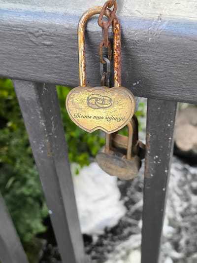 This media file captures a close-up of a heart-shaped padlock attached to a metal railing. The padlock is gold-toned and exhibits signs of rust, particularly on its chain and shackle. In the center of the padlock, there is an engraving of two intertwined rings, symbolizing a union or commitment. Below the rings, text is inscribed in a cursive font. The text reads: "Dievas mus sujungė."

Behind the padlock, out of focus, is a scene of lush green foliage and a body of water, likely a river or stream, with white, frothy water suggesting movement. The railing itself appears to be part of a bridge or a similar structure. The overall impression is one of a romantic gesture, possibly left by a couple to commemorate their relationship. The context provided indicates the location as Mosėdis, Lithuania. The lighting suggests it is daytime.