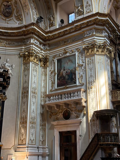 The image shows the opulent interior of a church in Mozzo, Italy. The architecture is characterized by a rich Baroque style, with white walls adorned with intricate gold detailing and ornate moldings. Sunlight streams in from a window on the right, illuminating the gilded capitals of columns and the decorative elements on the walls.
A large painting depicting a religious scene, with figures in classical attire and cherubs in the sky, hangs prominently on the wall above a decorative balcony. Below the painting, a door with a dark wooden frame is visible. To the right, a pulpit with a dark wooden structure and intricate carvings stands on a staircase. On the far left, a statue of a seated figure holding a severed head can be seen, with a painting of angels above it. The overall impression is one of grandeur and religious devotion.