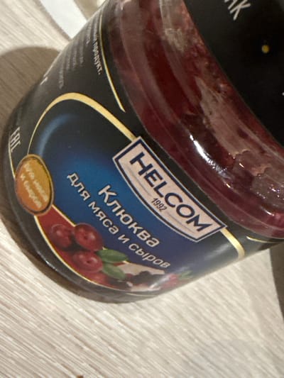 The image shows a close-up of a jar of cranberry sauce, likely "HELCOM" brand, as indicated by text on the label. The jar contains a dark red, viscous substance. The label is predominantly blue and black with gold accents. Prominent text on the label reads "КЛЮКВА" (Klyukva, meaning cranberry in Russian) and "для мяса и сыров" (for meat and cheeses). There is also an oval-shaped inset with text that is difficult to fully decipher but appears to be related to ingredients or use. The jar is placed on a light-colored, horizontally striped surface, resembling a wooden table or countertop. The lighting suggests an indoor setting, possibly with natural light creating shadows on the surface and highlighting the glossy texture of the jar. The scene is devoid of any contextual information related to Dubăsari, Moldova.