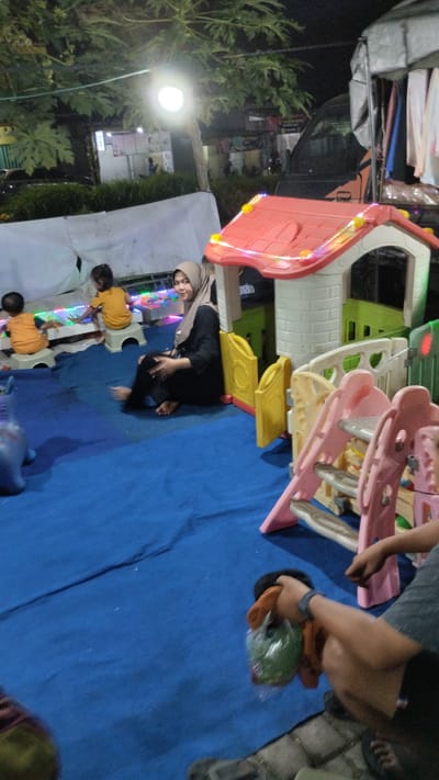 The image depicts an outdoor gathering at night, likely in Sukodono, Indonesia, indicated by the presence of a miniature playground structure with colorful lights. A woman wearing a hijab and black clothing is seated on the blue ground, looking towards the camera and smiling. In the background, two children in orange and yellow outfits are seated on small stools, engaged with illuminated toys or a play mat. A partially visible playground slide and fence are also present. To the right, a person's arm is visible, holding a plastic bag containing a green item. The environment appears to be a temporary setup, possibly a street event or a small amusement area, with a parked vehicle and a canopy in the background.