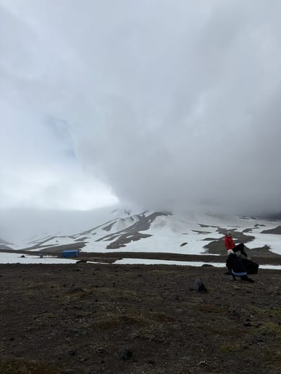 The image captures a person in a red jacket and white hat sitting on a rock in the foreground, with a snow-covered mountain shrouded in clouds in the background. A blue building and tents are visible in the mid-ground near the base of the mountain. The terrain in the foreground is dark and rocky. The scene appears to be an outdoor setting in a mountainous region, possibly a wilderness area near Petropavlovsk-Kamchatskiy, Russia, given the characteristic volcanic landscapes of the Kamchatka Peninsula.