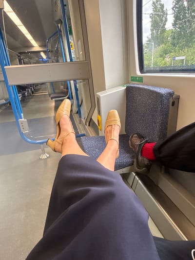 The image shows the interior of a train carriage, likely in motion, with views out of the window showing the blurred greenery. The window sign indicates the location is near Welwyn Garden City in Welwyn Hatfield, UK. Two people are visible: one with feet resting on a seat, wearing sneakers and red socks, and another with legs crossed, wearing tan-colored heeled shoes. The seats have a patterned blue fabric. There is a blue metal bar near the floor.