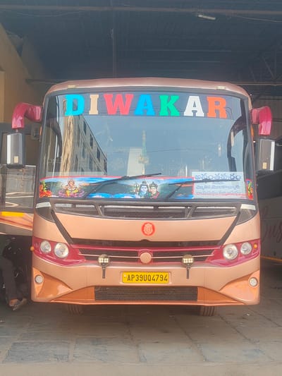 The image displays the front exterior of a gold and maroon colored 'DIWAKAR' passenger coach, registered under plate number AP39UQ4794, positioned under a covered structure. The bus is located at what appears to be a bus terminal or depot in Anantapur, India, as evidenced by a white route sign in Telugu script prominently displayed on the windshield, which clearly reads "అనంతపురం" (Anantapuram) and lists other destinations such as Chetlakor, Mulakalaluru, and Rayadurgam.

The bus features large colorful letters spelling "DIWAKAR" across the top of the windshield. Below the windshield wipers, a horizontal banner depicts Hindu deities, specifically Lord Ganesha on the left and Lord Shiva and Parvati on the right. A small, circular red emblem with a white Ganesha symbol is centered below the windshield. The front fascia includes dual circular headlights set within maroon bezels, accompanied by smaller circular LED lights beneath them. Two square auxiliary LED lamps are mounted centrally on the lower grille. The ground beneath the bus is paved with large, light-colored stone tiles. On the left side of the frame, the lower legs and feet of an individual, wearing dark trousers and light-colored sandals, are visible near the bus's front wheel. Reflections of an adjacent building are discernible in the bus windshield. A partial view of another vehicle or bus is visible to the right.