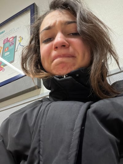 A young woman with brown hair and a black puffy jacket looks at the camera with a slight smile. She is close to the camera, and the image is taken at an angle. In the background, there's a framed advertisement with an illustration of people and app icons on a tablet. The wall behind the woman appears to be off-white.