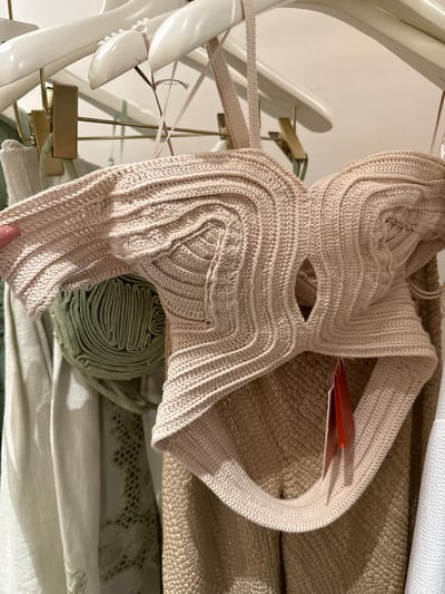 The image shows a beige knitted top on a hanger, hanging among other items in what appears to be a retail setting. The top has a unique, almost architectural design, with concentric rows of knitting creating a layered effect. A price tag is partially visible on the top. Next to the top is a roll of green fabric. Beneath those items are layers of other fabrics, including a cream-colored textured material, and white fabric. The image was taken at an indoor location in the city of Sunrise, United States of America.
