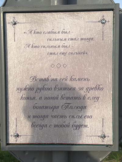 This image shows a weathered plaque mounted on a dark grey metal post in Cherdyn, Russia. The plaque is rectangular with ornate borders and four decorative corner elements, each containing a small screw. The text on the plaque is in Cyrillic script and appears to be an excerpt from a poem or proverb. The lighting suggests it is daytime, with shadows cast on the plaque from the post. The background is slightly out of focus but appears to be an outdoor setting with hints of green foliage. The plaque itself has a textured, metallic finish that shows some signs of wear and tear.