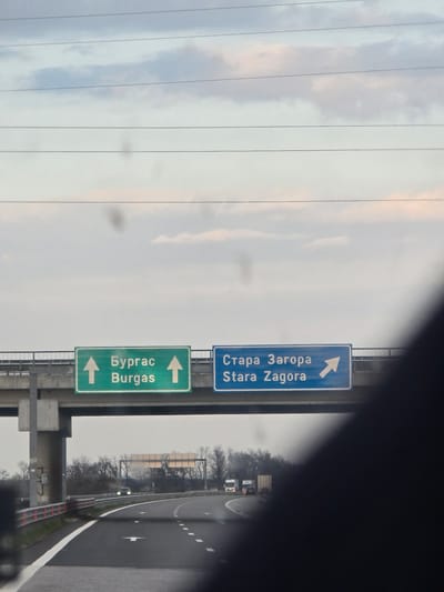 The image displays a view from inside a vehicle on an asphalt highway in Bulgaria, under an overcast sky. The scene is identified near Stara Zagora, Bulgaria, by prominent overhead road signs.
An overhead concrete overpass spans the highway. Below it, two rectangular road signs are affixed. The green sign on the left, marked with two upward arrows, displays "Бургас" (Burgas) and "Burgas," indicating a straight route. The blue sign on the right, with a diagonal upward-right arrow, displays "Стара Загора" (Stara Zagora) and "Stara Zagora," indicating an upcoming exit or turn-off. A thick, grey concrete pillar supports the overpass structure on the left side of the road.
The highway features at least two lanes of travel, demarcated by dashed white lines, with a solid white line defining the shoulder to the left. Reflective white arrows are painted on the road surface, guiding traffic. Red and white reflective guardrails line the left side of the highway. In the distance, the road curves to the right, where a light-colored vehicle with illuminated headlights is visible. Further right and partially obscured, two white trucks are present on the road. The surrounding environment consists of deciduous trees with bare branches, suggesting late autumn, winter, or early spring conditions. The sky is a uniform grey with subtle lighter patches, and several horizontal power lines are visible across the upper portion of the frame. Multiple dark, circular smudges are present on the viewing surface through which the photograph was taken. No individuals are visible. The implied action is vehicular travel.