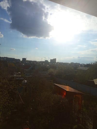 The image, originally rotated 90 degrees counter-clockwise, depicts a daytime landscape view overlooking a semi-urban area under a bright, partly cloudy sky. The sun, intensely bright and causing significant glare, is prominent in the upper left section of the sky. A large, dark cloud occupies the lower left sky, with lighter clouds scattered across the blue expanse.
In the midground and foreground, dense deciduous trees with green foliage are visible, indicating late spring or early summer. Partially obscured by the trees on the far right is a reddish-brown structure with a sloped, possibly translucent roof, resembling a gazebo or shed.
The background shows a sprawling settlement of various buildings, characteristic of a town or city, potentially совхоза "Воронежский" in Russia. Several tall construction cranes are discernible on the distant horizon, suggesting ongoing development.
The scene is strongly backlit by the sun, leading to underexposed details in the foreground and midground. No specific human activity is depicted. A distinct grid-like pattern is visible across the entire image, indicating it was captured through a screen, likely a window screen. No text is visible.