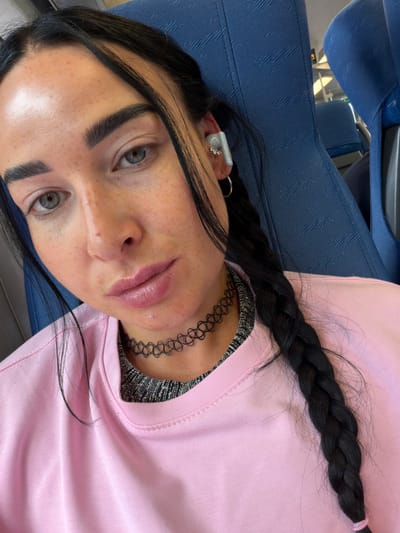 This media file shows a close-up, overhead shot of a young woman lying down on a blue seat, likely on public transportation given the texture and pattern of the seat. She is wearing a pink sweatshirt over a black and silver metallic top and a black choker necklace. Her long, black hair is styled in a braid that rests on her shoulder and the sweatshirt. She has striking light green eyes, dark eyebrows, and a scattering of freckles across her nose and cheeks. Her lips are full and appear to have a pinkish tint. She is wearing an ear cuff with small stones in her left ear. The lighting suggests it is daytime. The scene appears to be indoors, possibly in transit in or around Ушаки, Russia. There is no visible text in the image.