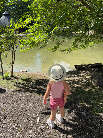 A young girl wearing a pink tank top, pink patterned shorts, a white straw hat with a cat face, and white sneakers stands facing away from the camera, looking towards a body of water. She is holding a doll. The ground is covered in gravel and scattered leaves. Behind the girl, a calm, green body of water reflects the sunlight and is partially shaded by overhanging tree branches with green leaves. In the water, two logs or debris are visible. To the left of the water, a small tree grows, and a dark-colored structure with a peaked roof, possibly a gazebo, is partially visible amongst the trees in the background, suggesting a park or recreational area. To the right of the water, a large tree stump is present. The scene appears to be outdoors in a natural setting.
