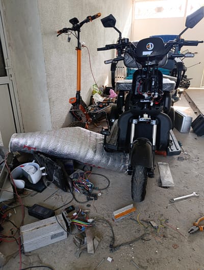 In Keŋ-Say, Kyrgyzstan, an outdoor concrete area adjacent to a building with a textured, light-colored wall and a partially visible white metal door shows two electric scooters and various disassembled electronic components. One scooter is orange and black, with its handlebars and front wheel visible. The other, a larger black scooter, is in the foreground and appears to be undergoing repairs or disassembly, with its front fairing removed, exposing internal wiring. Wires, circuit boards, a silver heat sink, a white plastic item with a handle, and a metal wrench are scattered on the ground around the scooters. A large, cylindrical object wrapped in reflective insulation material is also present. The scene suggests a workshop or storage area for electric vehicles and their parts.