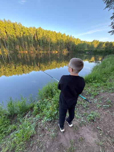 The image depicts an outdoor natural scene in the city of Mishkino, Russia, during daylight hours, likely morning or late afternoon given the quality of light. A young boy, viewed from the back, stands on a dirt path adjacent to a calm body of water, possibly a lake or large pond. He has short, light-colored hair and wears glasses with visible yellow temples. Dressed in a black long-sleeved shirt, black pants, and black and white athletic shoes, the boy holds a light blue and black fishing rod with both hands, its tip extended over the water. The water's surface exhibits a clear reflection of the sky and the opposite shoreline. The far bank is densely covered by a forest composed primarily of bright green deciduous trees, with some darker conifers interspersed. The near bank, where the boy stands, is characterized by green grass and various plants, transitioning to a visible dirt path. The sky above is clear and blue, with a few faint, wispy white clouds.