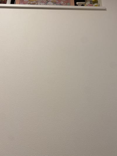 The image shows a white wall with a textured surface. The coordinates 34.693, 135.179 place the location in Kobe, Japan. The top edge of the wall is obscured by a shelf, out of frame, and the bottom edge of the wall is obscured by the bottom edge of the image frame. The wall is mostly white with a slightly off-white cast, and the texture appears to be a fine-grained, uniform pattern. There are no people, subjects, or objects visible in the image except the wall itself.