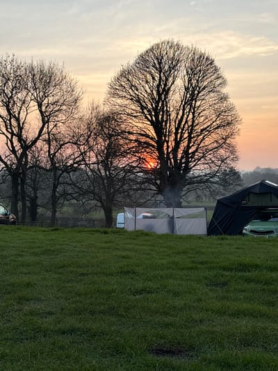 This image depicts an outdoor rural setting in Kirkby Malzeard, United Kingdom, during either sunrise or sunset. The sky displays hues of orange and pink at the horizon, transitioning to pale yellow and light blue-grey higher up, with the sun partially obscured by tree branches. The foreground consists of green grass, with a small dark depression visible in the lower center.
A row of bare, deciduous trees is silhouetted against the brightly lit horizon, dominating the mid-ground and background. A large, prominent tree is centered, with visible white smoke rising from its base, indicating a fire.
To the left, a portion of a light-brown vehicle is visible behind a wooden fence and trees. In the mid-ground, a grey, multi-paneled fabric windbreak is erected. A white vehicle, possibly a van, is partially visible behind this windbreak. To the right of the windbreak, a dark-colored tent or awning structure is set up, secured by yellow guy ropes. A light-green car or small SUV is parked beneath this structure, with a partial UK license plate reading "KX71" visible. A rooftop tent or storage unit is affixed to the green vehicle's roof. No individuals are present in the scene.