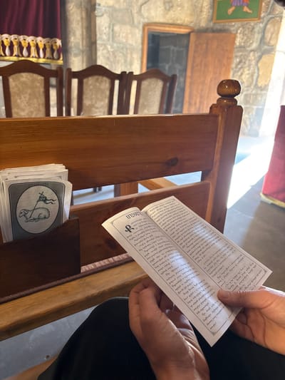 Inside a church, a person holds open a white booklet filled with text, seemingly reading or following along with a service. The book is held next to a wooden church pew. Inside a small compartment in the pew are stacked prayer cards with a drawing of a mammal holding a staff. In the background are other wooden pews, part of a stone wall, a piece of artwork, and a window. Sunlight streams in from above.