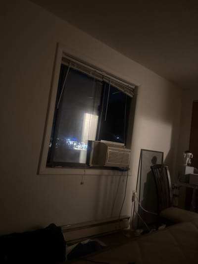 This is an indoor, eye-level, medium shot of a room at night. The room appears to be a bedroom, with a bed in the foreground covered by a dark blanket. To the left of the bed is a dark object, possibly clothing. In the background, a window shows a dimly lit cityscape, suggesting the location is the City of Binghamton, United States of America. The window has blinds that are partially open, and an air conditioner is installed in the lower part of the window frame. To the right of the window, there is a chair, a framed object leaning against the wall, and some medical equipment on a stand. The walls are painted white, and the lighting in the room is dim, with shadows cast by the objects.