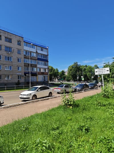 The image shows a street scene in Kumertau, Russia, on a sunny day with a clear blue sky. In the foreground, lush green grass and foliage line the side of a paved road. Several cars are parked or driving along the road. On the left side of the image, a multi-story apartment building made of light-colored bricks with blue trim is visible. The building has multiple windows, balconies, and air conditioning units. A street lamp stands near the building. On the right side of the road, a white signpost displays directions in Russian and English: "ГАФУРИ УР." (Gafuri St.), "ГАФУРИ УЛ." (Gafuri St.), and "GAFURI st." with arrows pointing left and right. Beyond the road, trees and other buildings can be seen. The overall impression is of a typical urban residential area on a bright summer day.