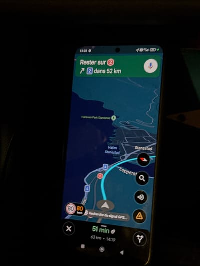 The image shows a smartphone screen displaying a navigation application. The time is 13:28. The navigation app indicates "Rester sur 2" (Stay on 2) for the next 52 km. A map of an area with a waterway and roads is visible. Text labels on the map include "Harissen Park Stansstad," "Hafen Stansstad," and "Stansstad." There are navigation icons indicating turns and route progress. A speed limit sign shows 80 km/h. Below the map, the display shows "51 min" and "63 km - 14:19." To the right of the screen, there are icons for search, volume, hazard warning, and a route icon. The background of the image is dark, suggesting the photo was taken in low light conditions, possibly inside a vehicle. The phone is held at an angle, with the screen facing the viewer.