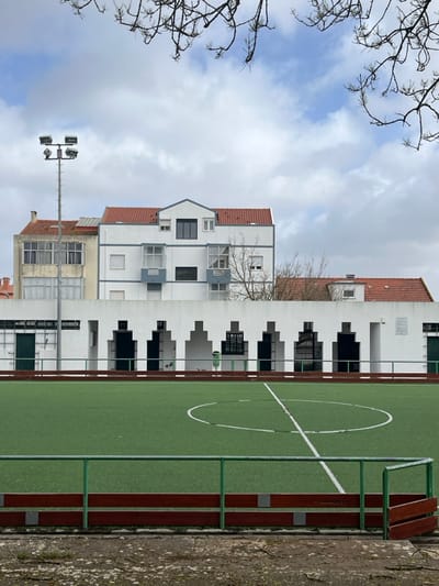 The image depicts an artificial turf sports field in an urban environment in Amadora, Portugal, under a partly cloudy sky. In the immediate foreground, a patch of barren ground transitions to a low, green-railed fence with a reddish-brown wooden kickboard. Beyond this fence lies a green artificial turf field, marked with white lines including a central dividing line and a central circle. No individuals are present on the field. To the left, a tall metal pole fitted with four floodlights extends upwards.

Behind the sports field and fence, a low, white architectural wall with a distinctive patterned facade features eight recesses or openings. Six of these are dark, suggesting entrances or deep shadows, while the leftmost two are partially obscured by a green metal door and a barred window. A green trash receptacle is visible within one of the recesses. A white access door is present on the far right of the wall, and a small white sign is affixed nearby.

Above and behind this white wall, several multi-story residential buildings are visible. The most prominent is a white building with a centrally gabled red tiled roof and grey-blue trim around windows and enclosed balconies. To its left, a lighter-colored building with multiple windows and enclosed balconies also features a red tiled roof. Further right and lower, another red tiled roof is visible. Bare tree branches are prominent against the sky in the upper frame, and some leafless trees are visible among the buildings.