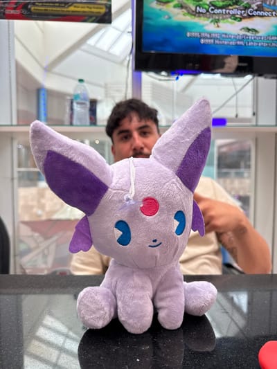A close-up, eye-level, medium shot captures a light purple stuffed toy, identified as Espeon from Pokémon, positioned on a black reflective countertop. The plush toy faces the viewer, with its large ears, pink and blue eyes, and small smiling mouth clearly visible. A white string with a clear plastic tag is attached to the toy's head. In the background, a man with dark hair and a beard is partially visible, out of focus, wearing a beige t-shirt. He is seated behind the countertop. Further in the background, shelves are stocked with various items, including a clear plastic water bottle, and a television screen displaying a colorful image and text, possibly from a video game. The environment appears to be an indoor retail or entertainment establishment, with the presence of a television and shelves suggesting a store or arcade. The image was taken in El Paso, Texas, United States of America.