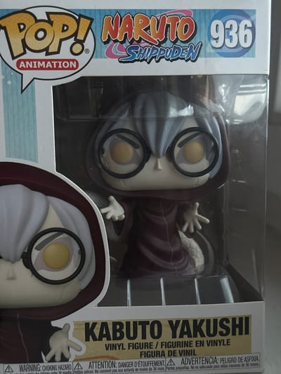 The image shows a Funko Pop! vinyl figure of Kabuto Yakushi from Naruto Shippuden, model number 936, in its original packaging. The figure is a stylized representation of the character, featuring his signature pale skin, round glasses, and dark hooded robe. The box has the Pop! Animation logo, the Naruto Shippuden title, and the model number clearly visible. The packaging is clear plastic, allowing the figure to be seen from the front. The bottom of the box displays warning labels in multiple languages, including "WARNING: CHOKING HAZARD" and "ATTENTION: DANGER D'ETOUFFEMENT". The background of the image is out of focus but appears to be a domestic setting. The image is a close-up shot of the product. There is no indication of the city of Żary, Poland, in the image.