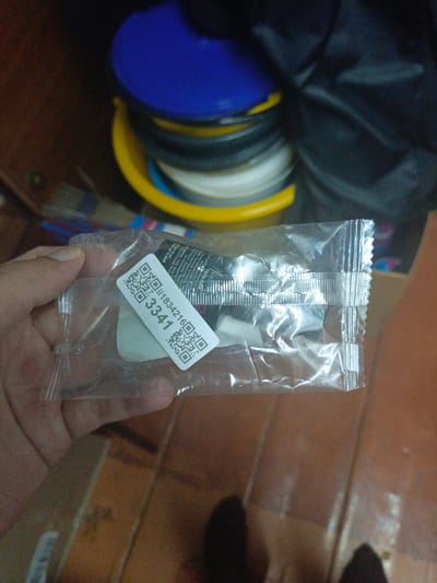 The image shows a person's hand holding a transparent plastic package containing a small white sticker with two QR codes and the text "ii1834216 3341". The package is sealed with a silver serrated edge. The background is out of focus, showing stacked colorful plastic lids (blue, yellow, and gray) and a black fabric object. The setting appears to be indoors with wooden flooring visible. The location of Ухманы, Russia is not ascertainable from the provided image.