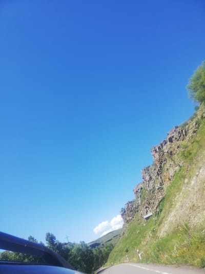 The image shows a scenic view of a mountainous landscape under a clear blue sky. A road curves through the foreground, with the perspective suggesting it is taken from a vehicle. To the right, a steep, rocky cliff face is covered in green vegetation, with scattered wildflowers. A small, box-like structure is visible built into the side of the cliff. In the distance, rolling hills and trees are present, along with a cross-shaped structure on a hill. The image is likely taken in the vicinity of Korkhi, Georgia, a region known for its natural beauty and mountainous terrain.
