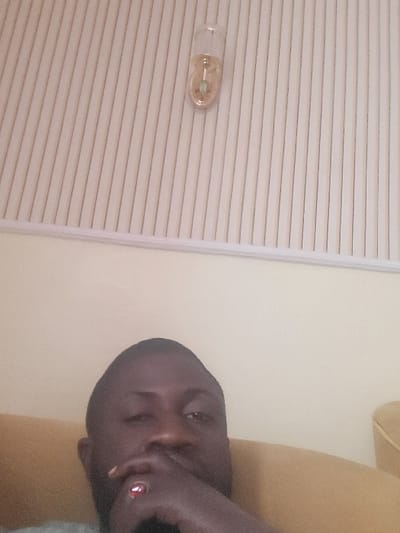 The scene is an indoor location in Babawa, Nigeria. A dark-skinned male is visible from the eyes down, occupying the lower half of the frame and looking directly forward. His right hand covers his mouth and chin, with a prominent red-stoned ring on his right ring finger. He is seated on light brown upholstered furniture, a segment of which is visible on the right. The upper background features light cream-colored vertical blinds or paneling. A small, transparent, cylindrical light fixture containing a green element is attached to this paneling in the upper right quadrant. A plain, light yellow wall is visible to the right of the blinds.