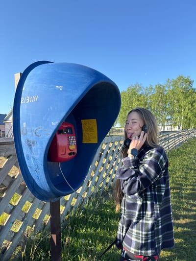 In Ust'-Silayka, Russia, a young Caucasian woman is captured in an outdoor scene on a clear, sunny day. She stands beside a freestanding blue public payphone booth, which is mounted on a dark metal pole. The woman has long, light brown hair, visible freckles, and wears a dark blue, green, and white plaid fleece jacket over dark pants patterned with red chili peppers. A black smartwatch with a white band is on her left wrist. She holds a black telephone receiver to her left ear with her right hand, simultaneously sticking her tongue out playfully.

The blue payphone booth, showing signs of wear with scratches, faded paint, and graffiti including the numbers "2131111" and a heart shape, houses a red payphone unit. The red unit features a numeric keypad and Cyrillic text, including "ФЕДЕРАЛЬНЫЕ УСЛУГИ СВЯЗИ РОССИИ" (Federal Communication Services of Russia). A yellow sheet of paper with black Cyrillic text is affixed to the interior right side of the booth. The receiver is connected to the unit by a coiled metal cord.

Behind the payphone and the woman, a light-colored wooden lattice fence extends across the mid-ground. Beyond the fence, green grass with dandelions covers the ground. A line of birch trees with bright green leaves and white trunks is visible in the background, along with several distant buildings, including a light-colored wooden house with a light blue roof and a larger white structure. The sky is clear and bright blue, featuring a faint crescent moon in the upper right quadrant. Strong sunlight illuminates the scene, casting distinct shadows.
