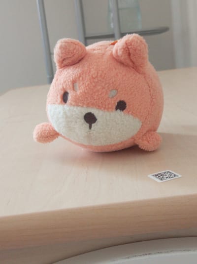 A fuzzy, peach-colored plush toy resembling a fox or dog sits on a light-colored wooden surface. The toy has round ears, two dark oval eyes, a small dark nose, and a white muzzle with two light-colored oval cheeks. One of its paws is extended to the left. To the right of the toy, a small, white, square sticker with a QR code is visible. The background consists of a blurry light gray wall and vertical supports. The scene appears to be indoors, on a table. There are no people present. The location in Chapurniki, Russia, is not discernable from the image.