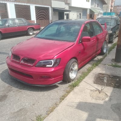 A modified dark red sedan, identifiable as a Mitsubishi Lancer, is parked diagonally on a street in Barquisimeto, Venezuela. The vehicle occupies both a portion of the asphalt road and the adjacent cracked concrete sidewalk. It features a black roof, aftermarket headlights with integrated LED daytime running lights, a custom front bumper with a lower lip, and polished multi-spoke wheels with stretched tires. A rear spoiler is visible. The hood appears slightly ajar on the passenger side. To the left, an older two-tone black and red sedan is parked on the street. Behind the red Lancer, a dark-colored SUV or wagon is partially visible. The background consists of residential buildings with light-colored facades, some featuring security grates or roller blinds over windows. A dark utility pole with a metal chain attached to the ground stands on the right side of the sidewalk. Weeds are visible growing through cracks in the concrete and along the curb. The scene is illuminated by daylight, casting discernible shadows. No individuals are present.