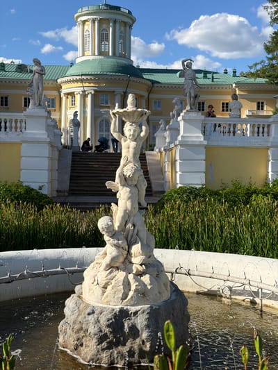 This media file depicts a scene at Arkhangel'skoe, Russia, featuring a grand yellow building with white architectural details, including columns, balconies, and a dome. In the foreground, a water fountain with intricate stone sculptures of cherubic figures is active, with water spraying and falling into the basin. Lush green grass and hedges surround the fountain and the building.
Several people are visible in the scene, some standing on the balconies of the building and others on the steps leading up to it. One person is seen near the fountain, observing the sculptures. The weather appears to be clear and sunny, as indicated by the bright lighting and the presence of scattered white clouds in the blue sky. The time of day seems to be daytime, likely during sunny hours.
Notable details include the classical style of the architecture and the detailed craftsmanship of the fountain sculptures. The composition of the image, with the fountain in the foreground and the impressive building in the background, creates a sense of depth and scale. The overall mood conveyed is one of serene beauty and historical grandeur. No visible text is present in the media file.