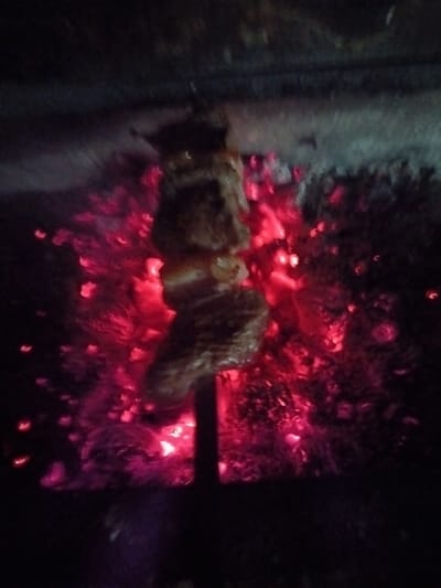 Skewered meat cooks over red-hot embers in a dark setting. The meat appears to be in three distinct pieces, slightly charred. The intense heat emanates from the smoldering coals surrounding the skewer, creating a fiery glow. The image is dimly lit, with the focus on the cooking process and the vibrant embers.