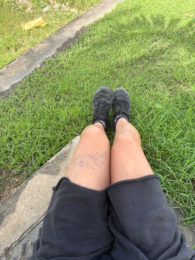 In Tambon Hua Toei, Thailand, the image presents an overhead view of a person's legs and feet. The subject is wearing black shorts, black athletic socks with white accents, and black, worn athletic shoes. A black outline tattoo resembling a stylized marine creature or fish skeleton is visible on the front of the left thigh. The legs are positioned over a predominantly green grassy area. A concrete curb or edge is present in the foreground and diagonally traverses the upper-left portion of the frame. A crumpled yellow plastic object lies on the grass near the concrete curb in the upper-left. The scene is illuminated by natural light.
