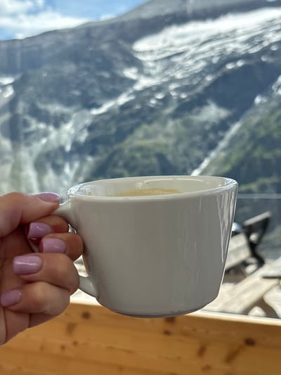 A white coffee cup, held by a person's hand with light purple-painted fingernails, is in the foreground. The cup contains a light-brown liquid, possibly coffee. In the background, out of focus, is a mountainous landscape with snow-capped peaks and rocky slopes. The sky is partly cloudy. The scene appears to be set on a wooden surface, possibly a table or balcony railing. The overall impression is of enjoying a hot beverage with a scenic mountain view, likely in Heiligenblut am Großglockner, Austria, given the presence of the Großglockner mountain, which is Austria's highest mountain.