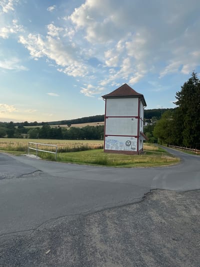 The image depicts a view of a rural landscape near Neukirchen, Germany, featuring a distinctive square tower building. The tower is white with red trim along its edges and roofline, and a red tiled roof. The lower portion of the tower is adorned with graffiti or murals. The building stands on a grassy area, with a paved road curving around it. In the background, there are fields, trees, and a wooded hill. The sky is blue with scattered white clouds. A metal railing is visible in the foreground on the left, along the edge of a field. A wooden fence runs along the right side of the road in the background. The overall scene suggests an outdoor, natural setting with a man-made structure integrated into the environment.
