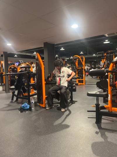 A person in a white soccer jersey with the Palestinian flag and the word "فلسطين" in Arabic is working out at a gym. They are using a seated row machine, with their legs extended and hands gripping the handles. The gym is brightly lit with orange and black equipment. A water bottle and towel sit on the machine next to them. Other gym equipment and people are visible in the background.