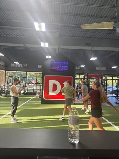 The image depicts the interior of a brightly lit gym, likely a D1 Training facility, given the prominent "D1" logo. The gym features a turf floor with white lane markings and various exercise equipment, including treadmills and weight racks, visible in the background. Natural light streams in from large windows, illuminating the space.

Several individuals are engaged in physical activity. In the foreground, a man wearing a white tank top and dark pants is standing and looking at his phone. Further back, a group of young people are participating in a workout. A blonde woman in a maroon shirt and black shorts is in mid-stride, her arms outstretched. To her left, a boy in a khaki shirt and grey shorts is also in motion. Another boy in a grey t-shirt and grey shorts is further back. A person in a patterned shirt is seated on the turf.

The gym's walls are adorned with motivational words like "DETERMINATION," "DISCIPLINE," "INTEGRITY," "NATION," and "HONOR." A large screen displays a colorful animated scene. A clear plastic water bottle sits on a dark surface in the foreground. The ceiling is high, with exposed metal beams and industrial lighting fixtures. The overall impression is a dynamic and active fitness environment.