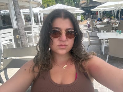 A woman is taking a selfie in Key Largo, Florida, USA. She is wearing a brown ribbed tank top and round, gold-rimmed sunglasses. She has a gold necklace. Her hair is curly and wet. She appears to be sitting at an outdoor restaurant or bar. There are white chairs and tables around her, as well as umbrellas. In the background, people can be seen dining and socializing. Palm trees are also visible.
