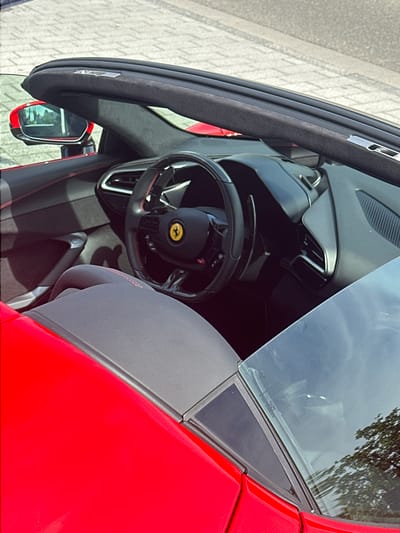The image depicts the interior of a red Ferrari sports car. The steering wheel, with a yellow Ferrari emblem, is visible. The driver-side door is ajar, revealing the car's black interior. The environment outside the car shows signs of Jestetten, Germany, with light-colored pavement visible in the background. The windshield reflects foliage, suggesting an outdoor setting.