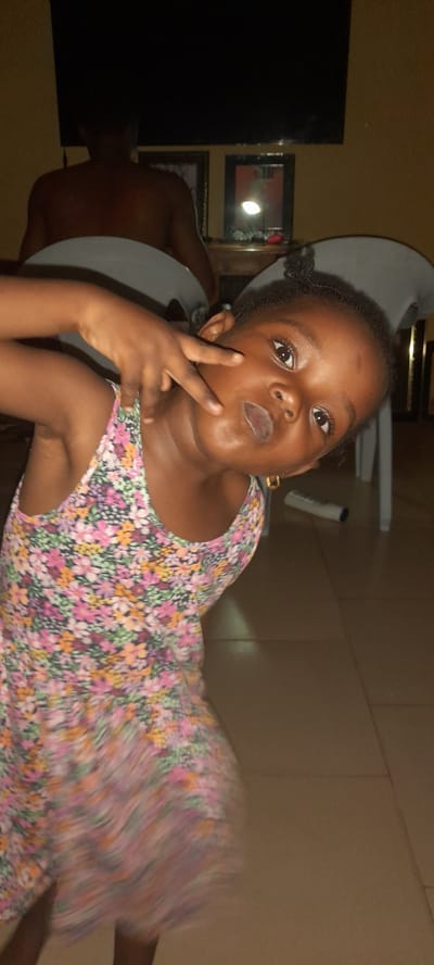 A young girl, possibly four to six years old, is lying on her side indoors. She is dark-skinned and wearing a floral-patterned dress with a pink, orange, and green floral print. Her hair is styled in small braids. She is holding her right hand up, making a peace sign near her cheek.

In the background, there is a dark, possibly black, television or mirror. A person is sitting behind the girl wearing a grey shirt. The background wall appears to be painted in tan. The location is Ashipa, Nigeria.